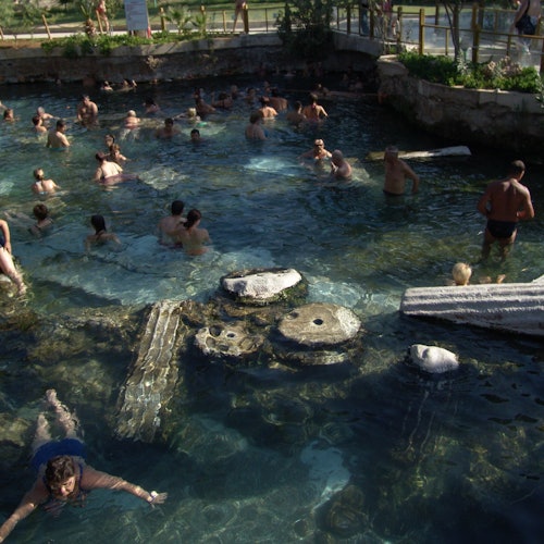 Mga tao na lumalangoy sa isang natural na pool na may bahagyang nakalubog na sinaunang mga haligi at bato. Ang lugar ay napapaligiran ng luntiang mga halaman.