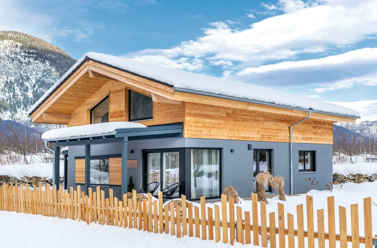 Stilvolles Holzhaus mit grauer Fassade im Winter, bedeckt mit Schnee, vor bergiger Kulisse.