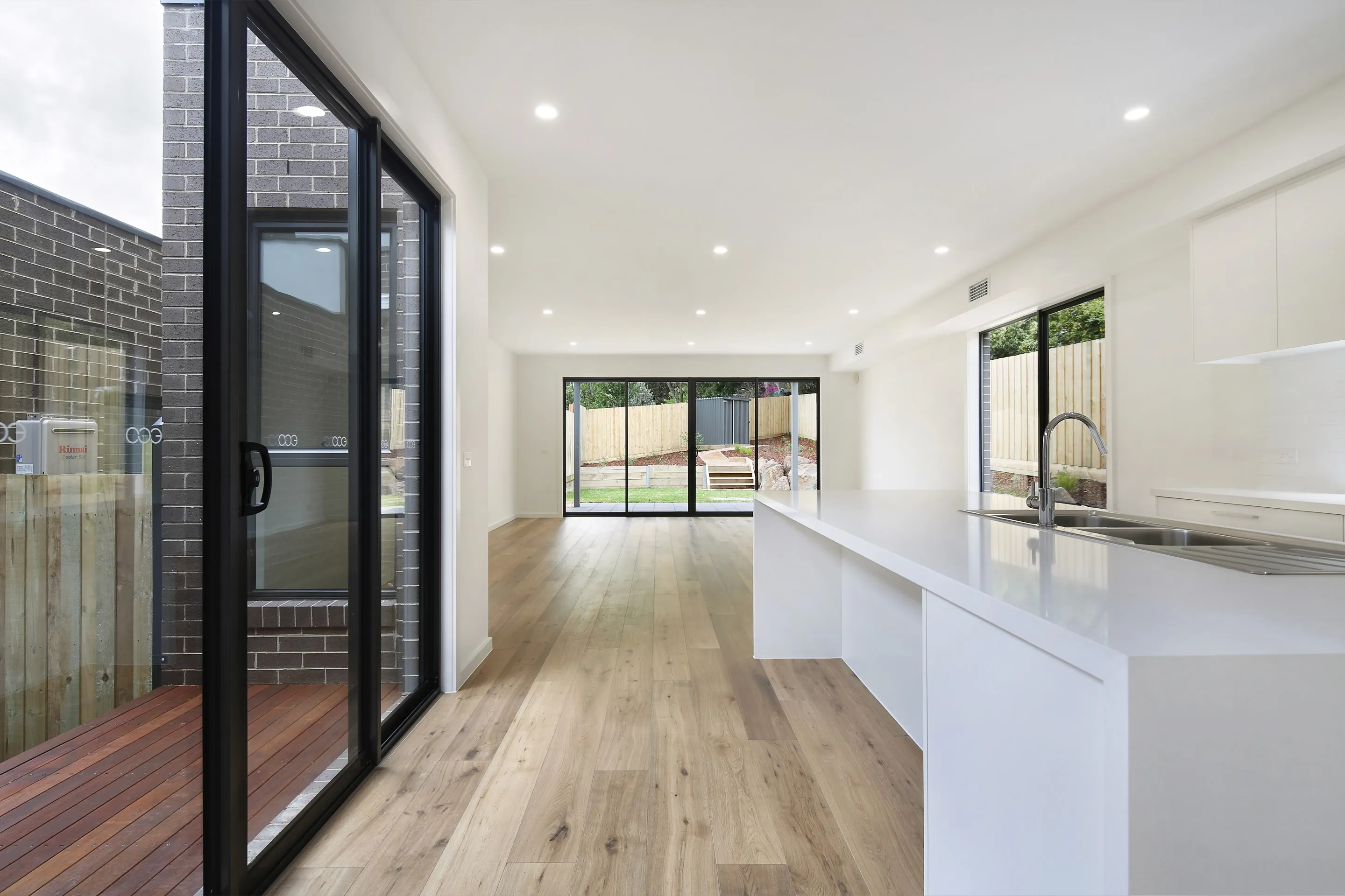 Open living looking through black-framed sliding doors to a landscaped backyard, with a white waterfall island and sink, a brick light court and a Rinnai unit on the wall
