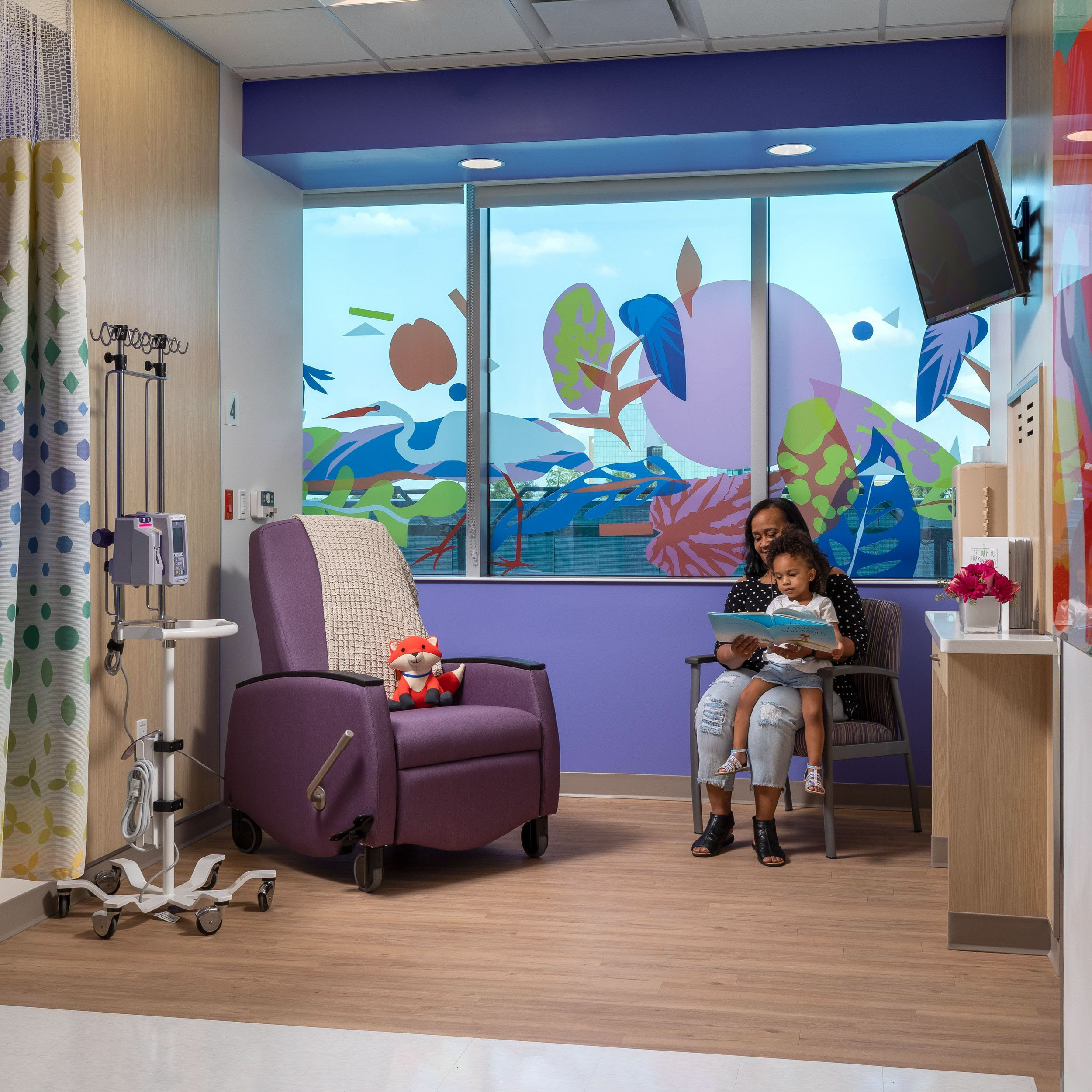 Woman reads book to child in a colorful hospital room.