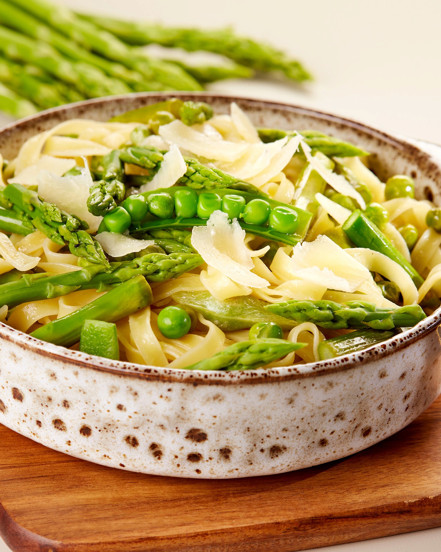 A rustic ceramic bowl filled with tagliatelle pasta, topped with fresh green asparagus, sweet peas, and shaved Parmesan cheese, arranged on a wooden table for a vibrant spring dish.