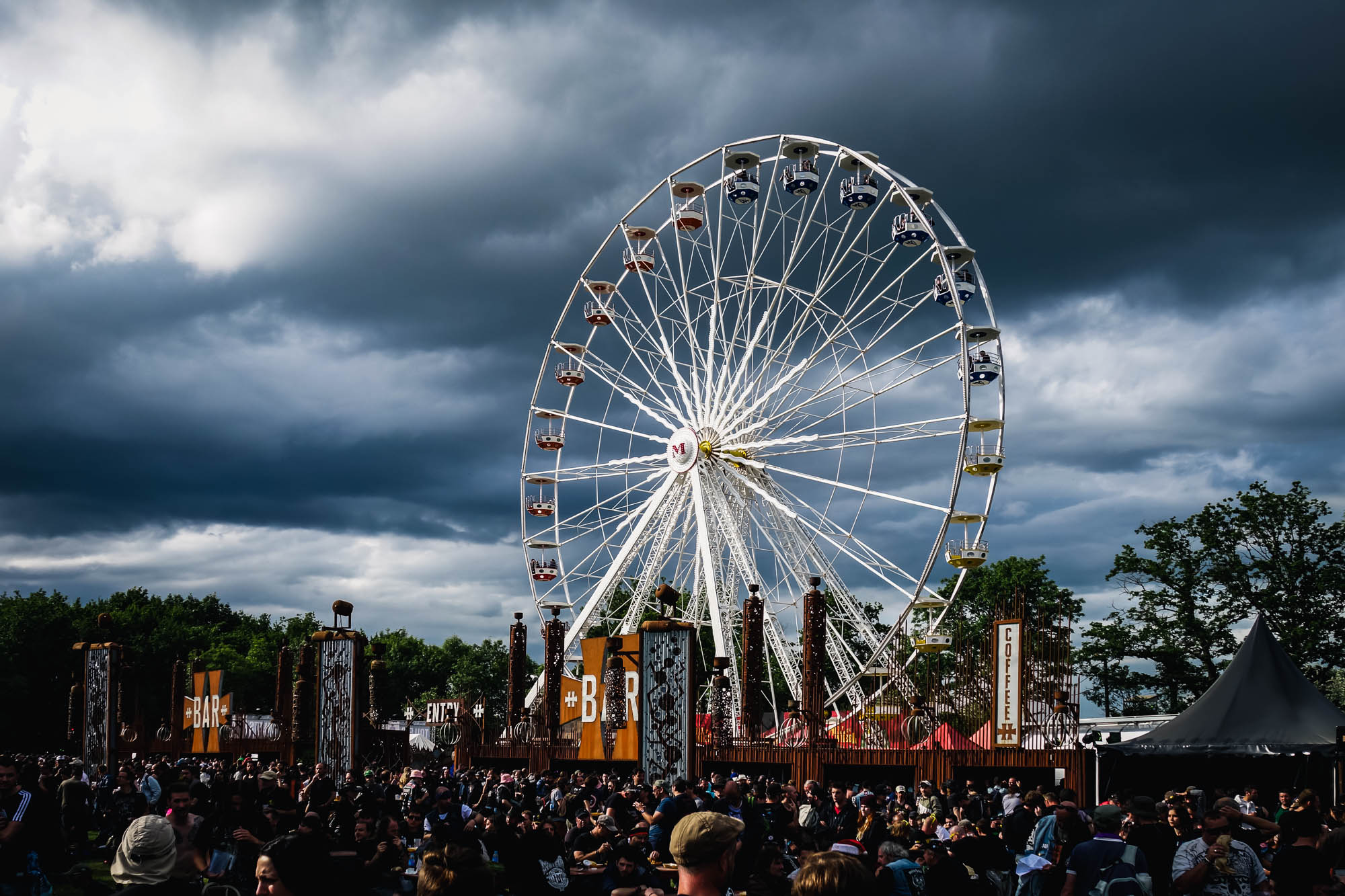 Grande roue du festival sous un ciel d'orage, foule assise au premier plan, Hellfest
