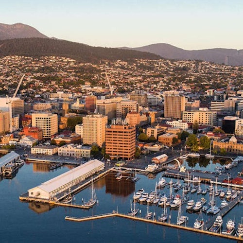 Въздушен изглед на крайбрежен град с лодки, акостирали на пристанище, разнообразни сгради и хълмове на заден план.