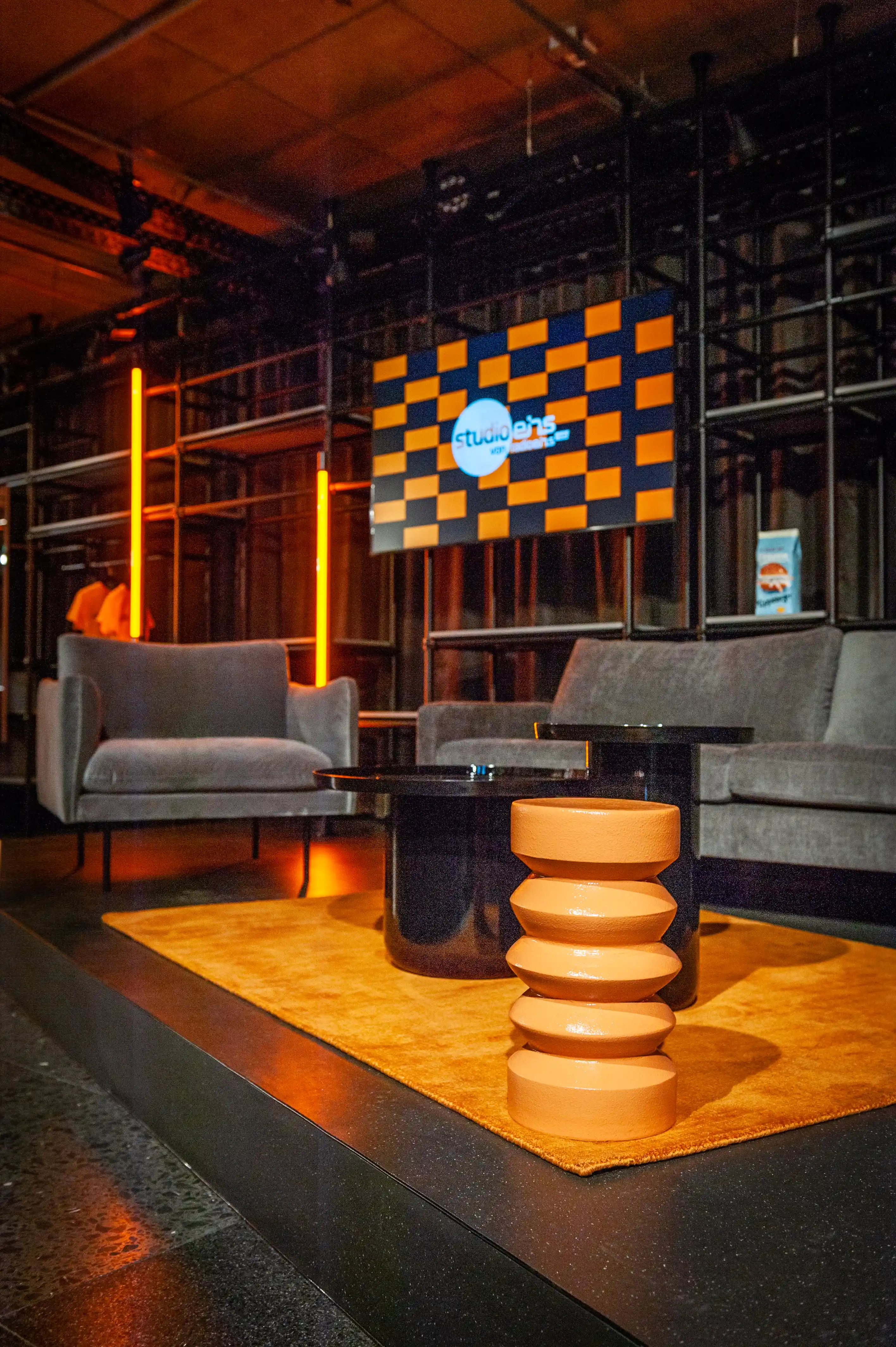 Cozy lounge in the Radio Eins studio with grey armchair, sculptural terracotta stool and glowing screen in checker design.