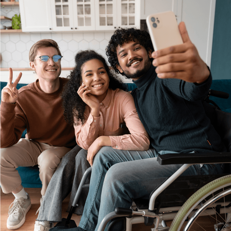 Three friends, one in a wheelchair, sit together on a couch smiling and posing for a selfie. One person makes a peace sign while another touches her face. They appear happy and relaxed in a bright, modern living room.