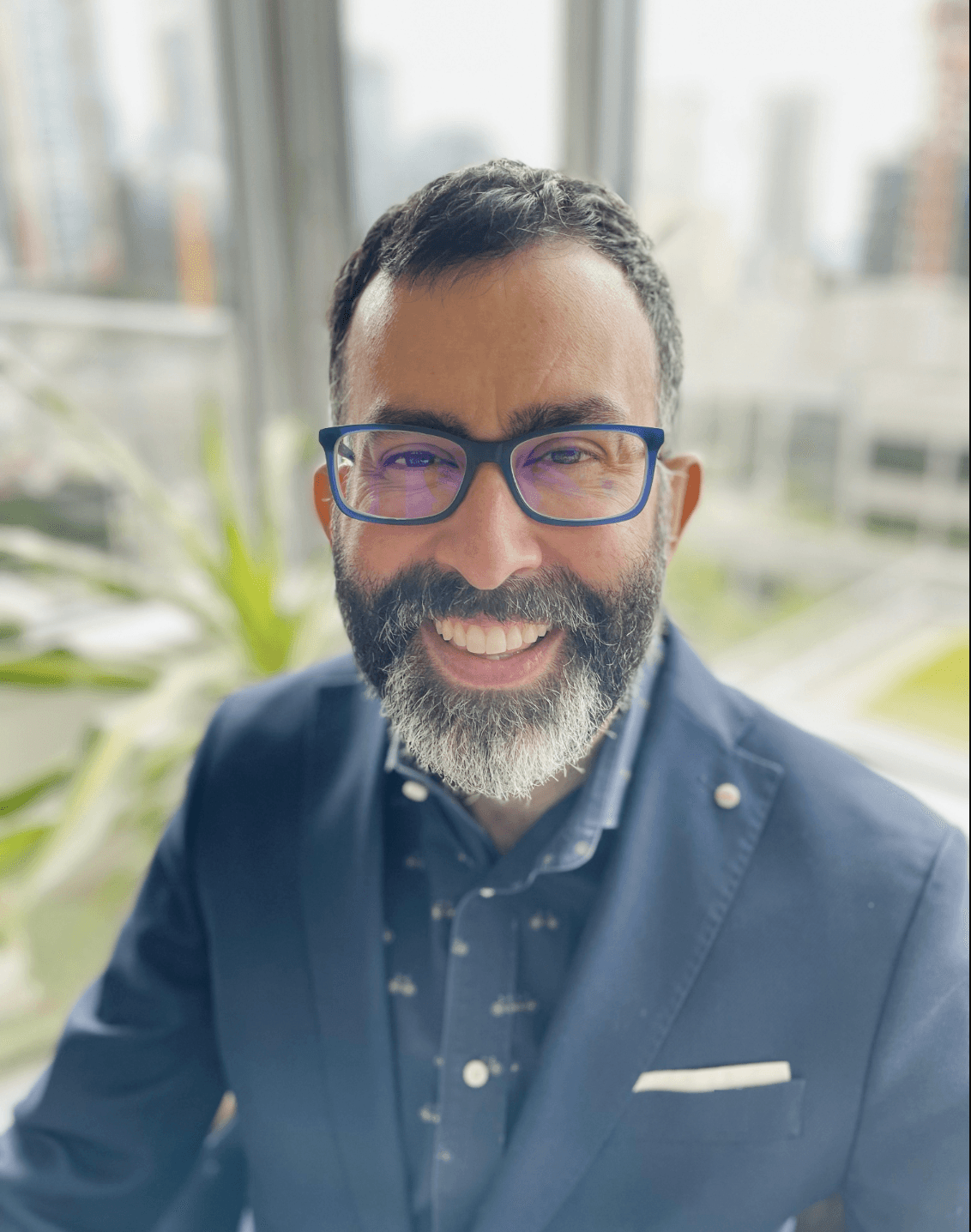 A smiling man with a beard and glasses, wearing a navy blazer, sits indoors with city buildings in the background.