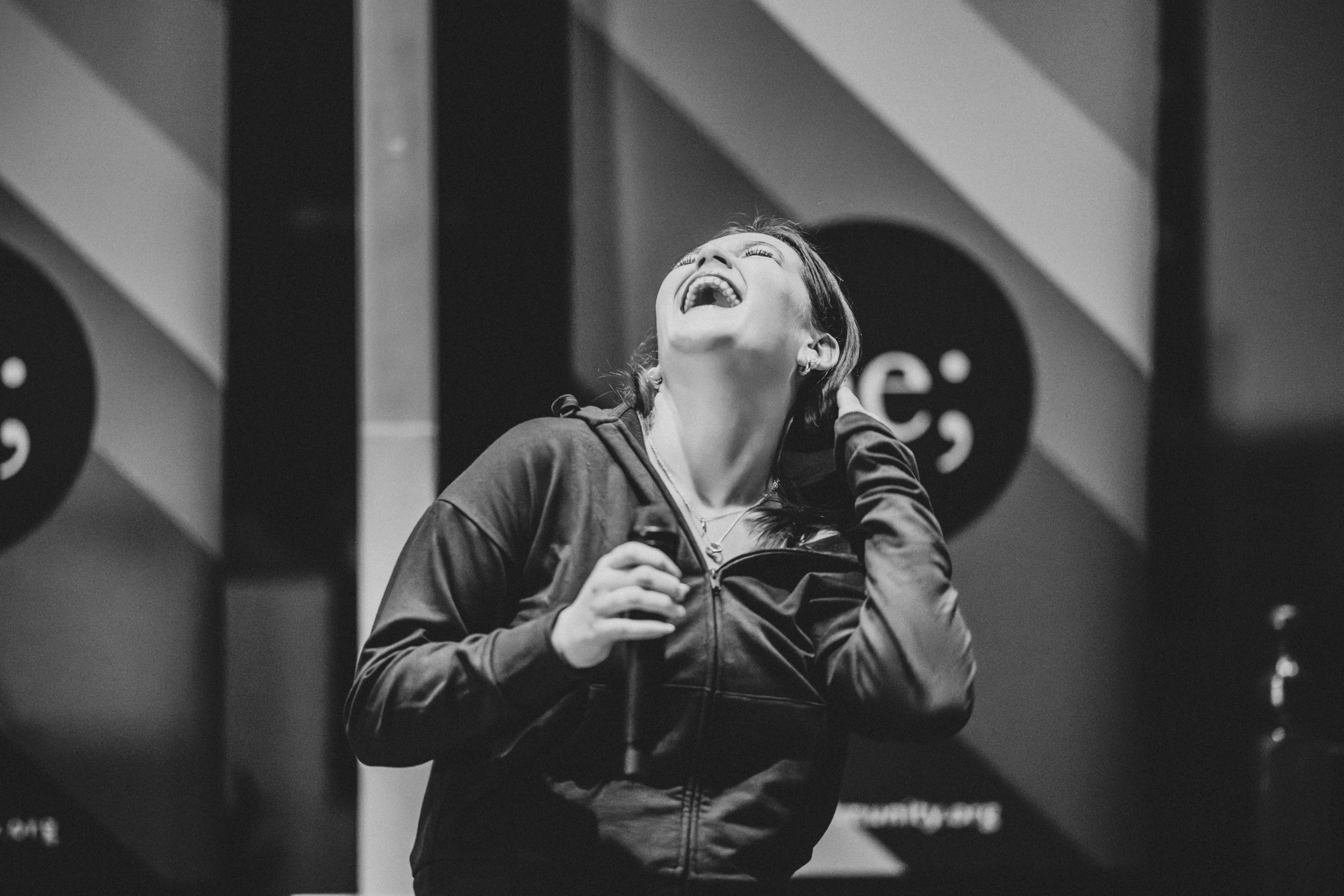 Performer laughing mid-performance while holding a microphone at the Queer Open Mic (QOM) hosted by be; community in Bridgewater, Massachusetts