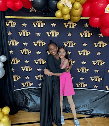 Two teens hug in front of a VIP step-and-repeat backdrop that looks like a movie red carpet.