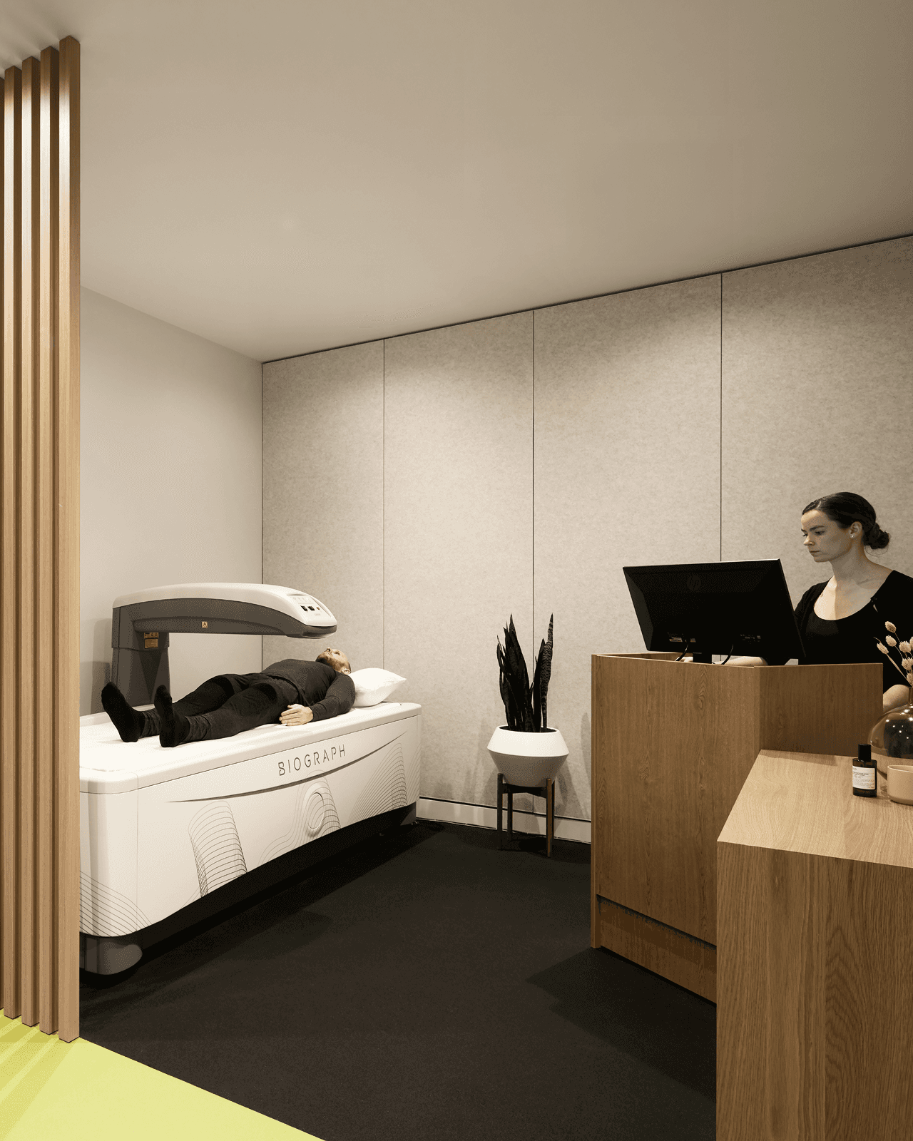 Patient lying on a diagnostic scanning table in a private clinic room while a technician works at a computer station.