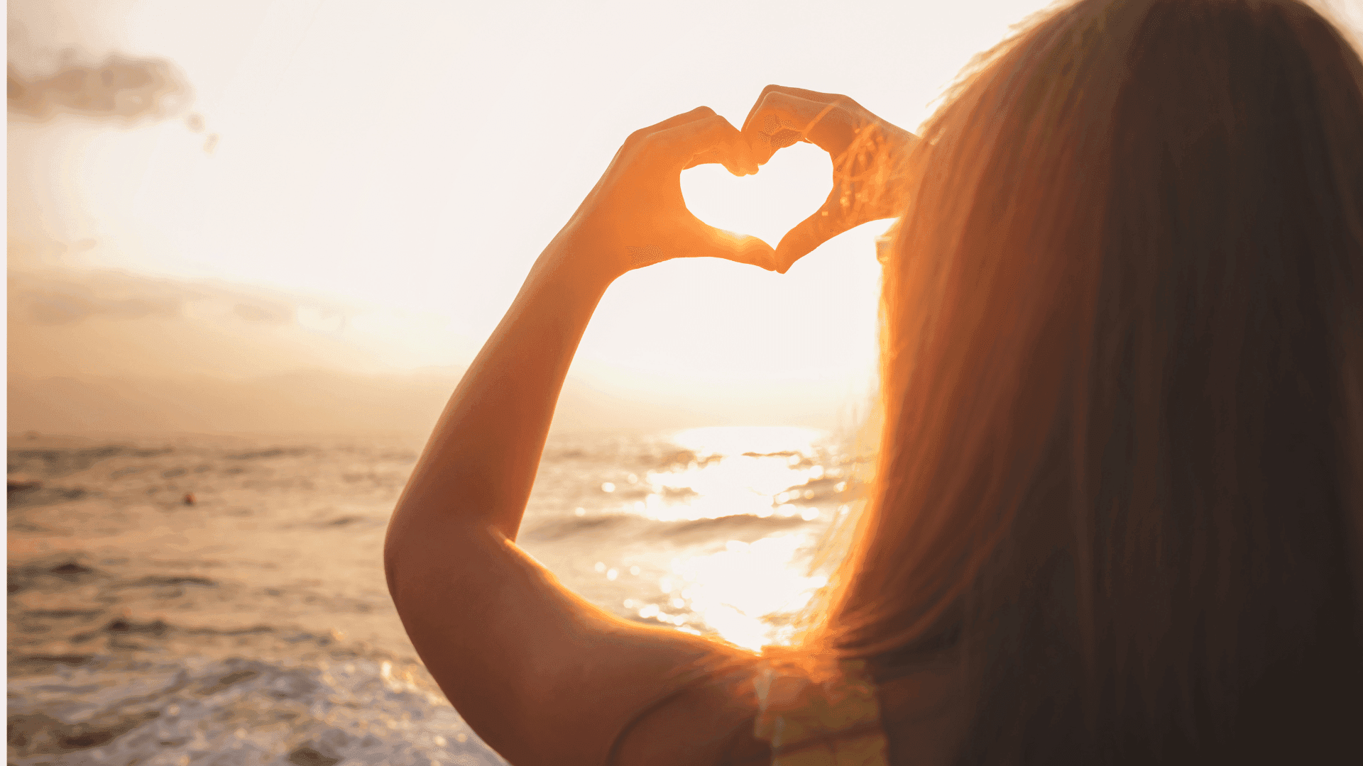 A woman on the beach making a heart shape with her hands, with the sun shining through the centre of the heart.