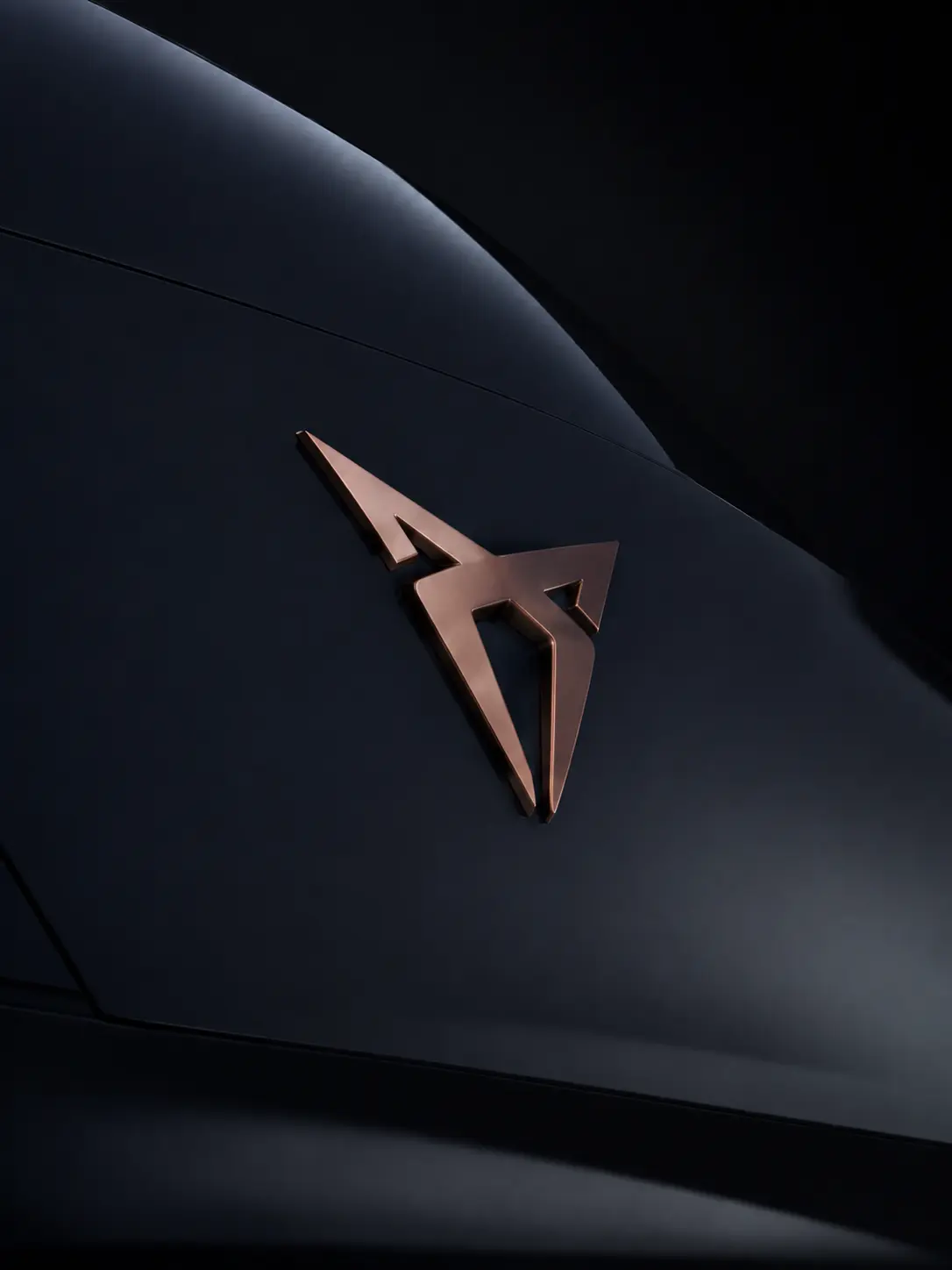 Kupferfarbenes Cupra-Emblem auf einer dunkelblauen, matt lackierten Fahrzeugkarosserie.
