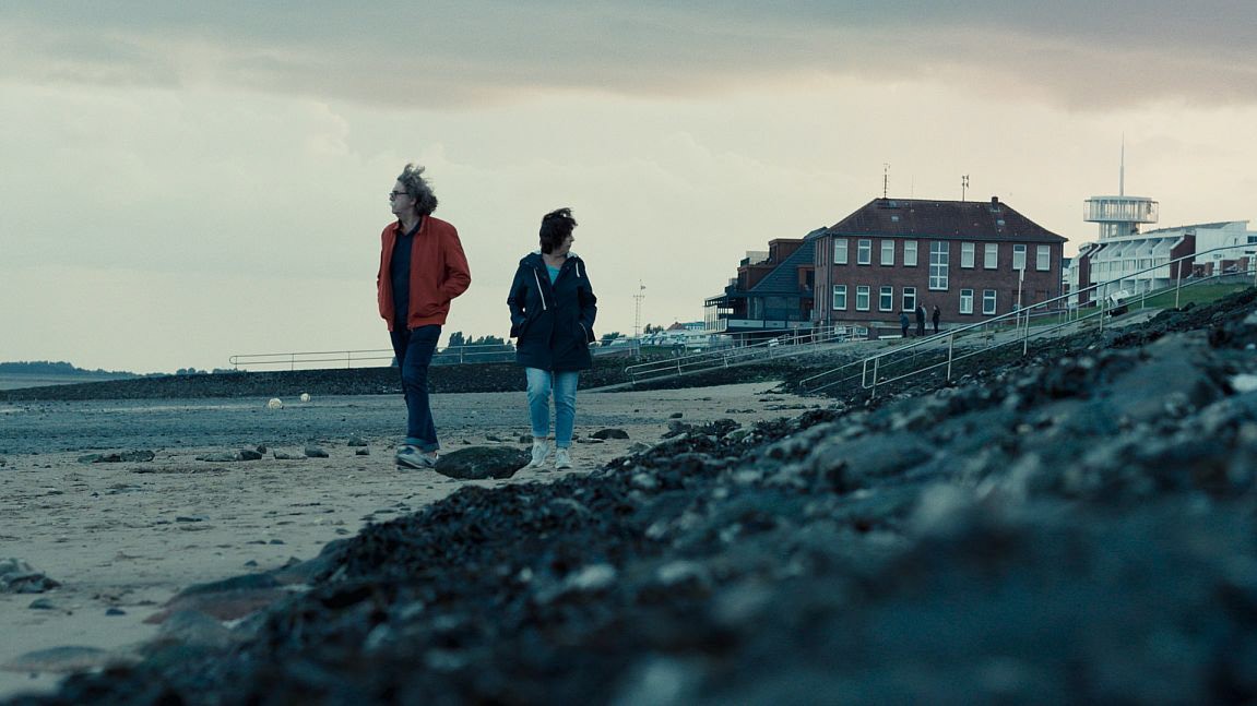 Two figures stand on a beach, looking out towards the ocean under a cloudy sky. A distant building is visible.