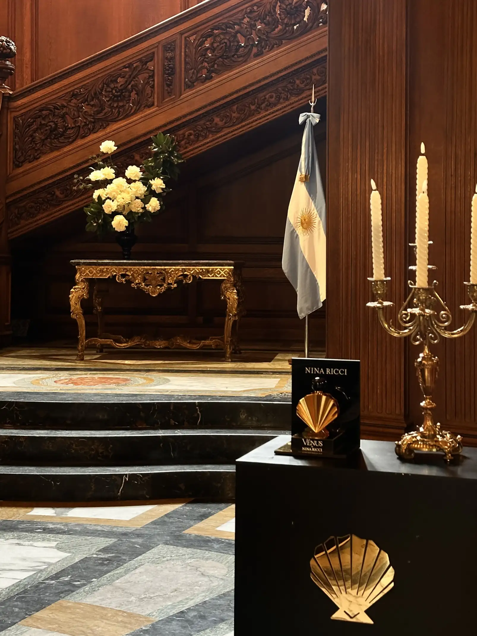 Vestíbulo elegante con escalera de madera tallada, mesa dorada con arreglo floral de flores blancas, bandera argentina colgada junto a la pared y candelabros dorados; en primer plano, un pedestal negro con el perfume Venus de Nina Ricci exhibido.