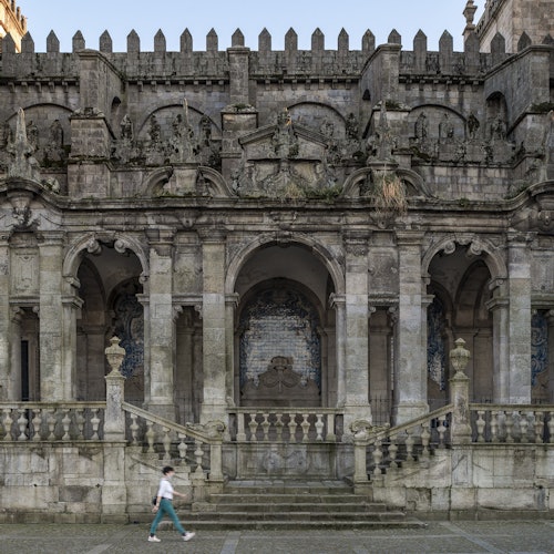 A person walks in front of an ornate, old stone building with arched entryways and detailed architectural carvings.