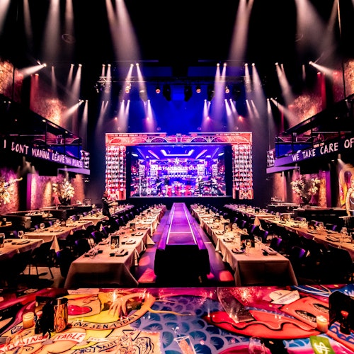 Sebuah aula makan yang hidup dan penuh warna yang disiapkan untuk sebuah acara, dengan meja panjang mengarah ke panggung yang diterangi dan dihiasi dengan lampu neon dan lampu sorot.