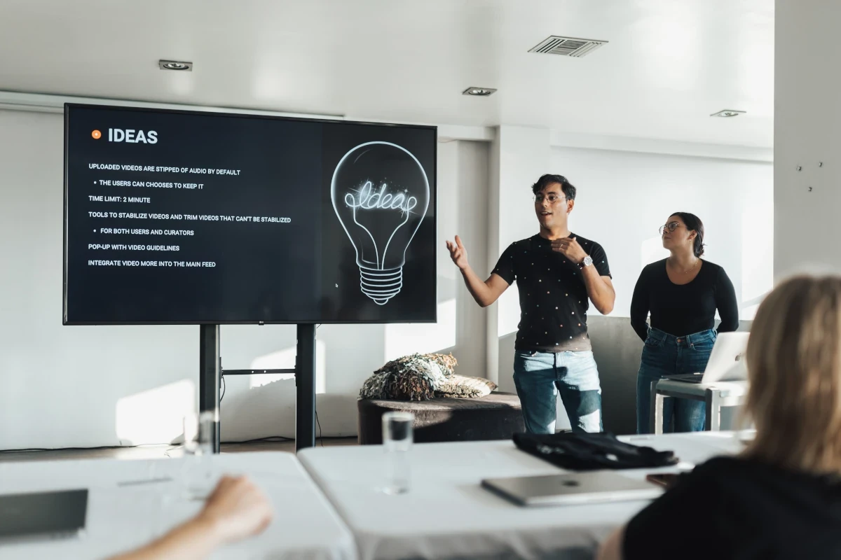 Man giving a presentation beside a large screen, speaking confidently to a group.