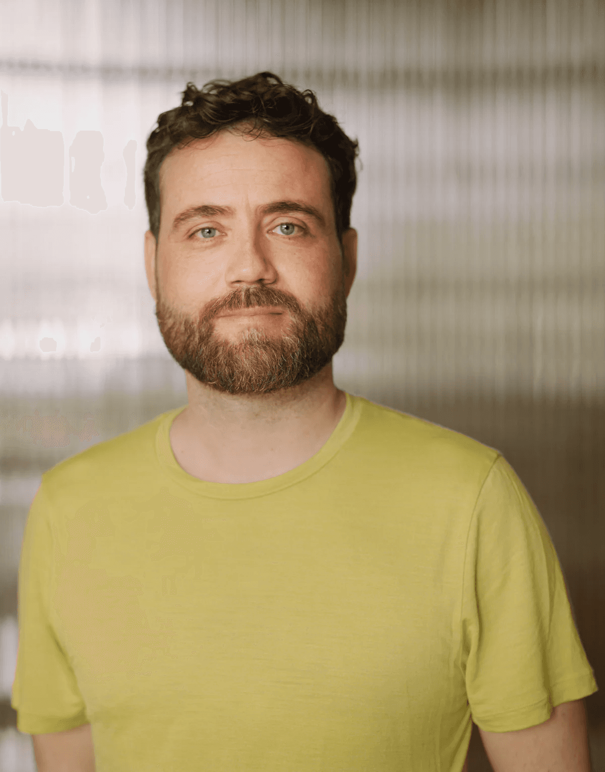 A man with a beard and curly hair is wearing a light green T-shirt, standing in front of a softly blurred, striped background.