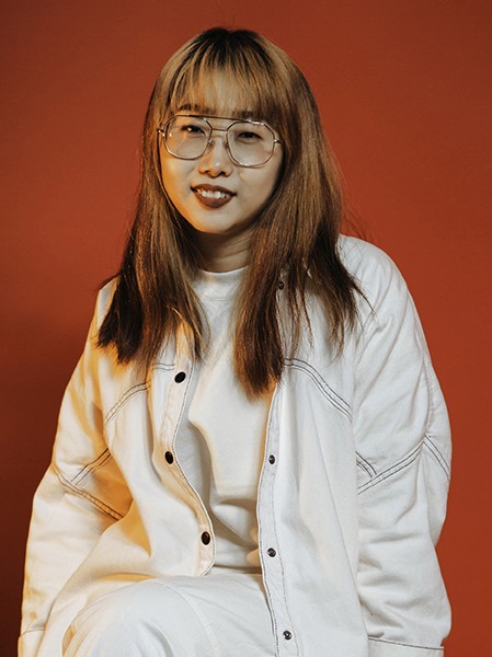 A person with shoulder-length brown hair with bangs and thin-framed glasses sits against a solid orange-brown background. They are wearing a white long-sleeved shirt with black buttons and black trim on the sleeves and collar. The shirt is open at the front, revealing a white undershirt. The person’s expression is neutral, and they are looking directly at the camera.