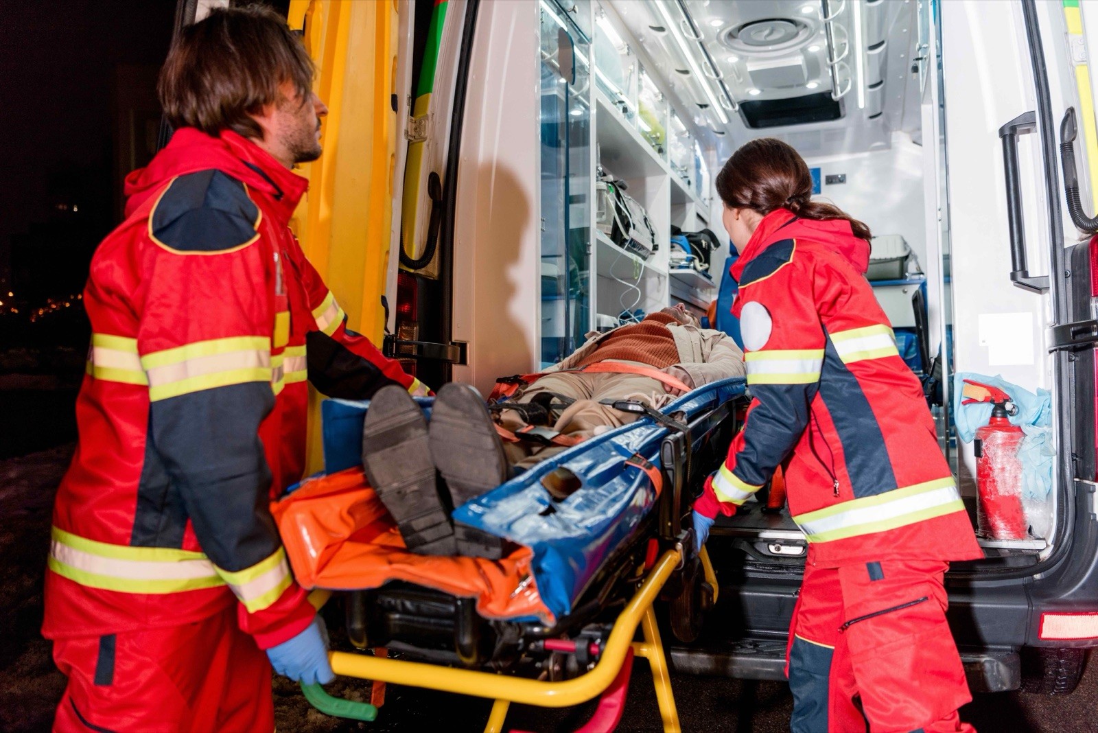 Two paramedics in red uniforms carefully load a patient on a stretcher into the back of an ambulance.