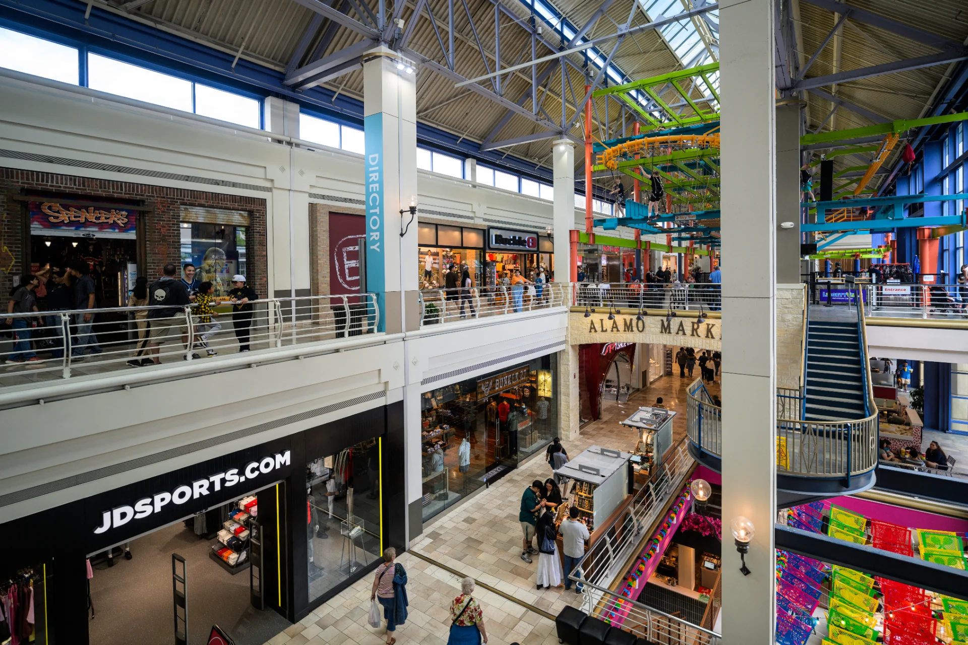 Multi-level interior of the Shops at Rivercenter showing JD Sports and Spencer's storefronts, Alamo Market Place, an overhead ropes course, and shoppers on multiple floors
