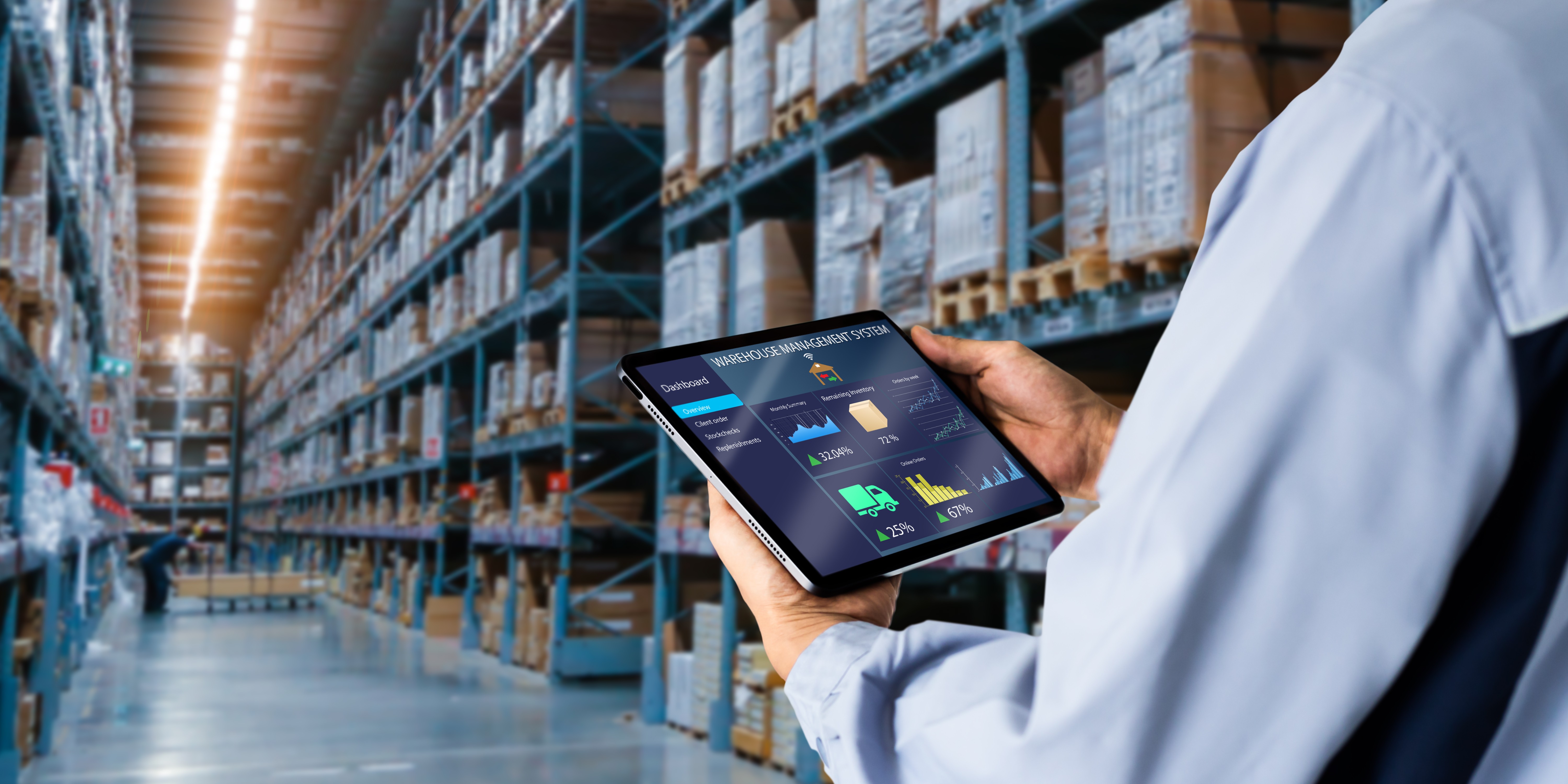 A person uses a tablet to monitor inventory analytics in a large warehouse, with rows of shelves filled with stacked boxes extending into the distance, illustrating modern logistics and supply chain management.