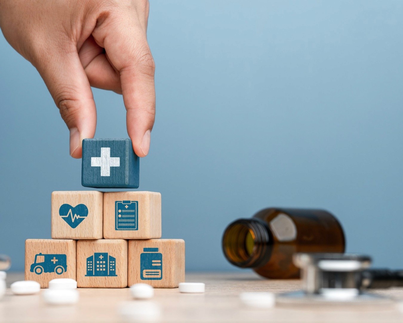 A hand places a block with a medical cross on top of stacked blocks with healthcare icons: heart, clipboard, ambulance, hospital, pills.