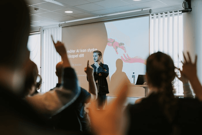 A speaker stands at the front of a room, while attendees raise their hands for questions or participation.
