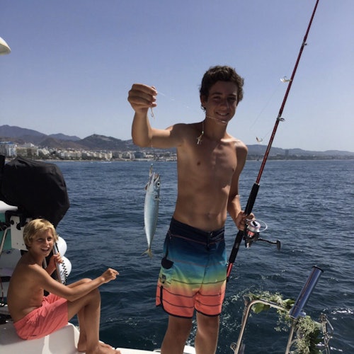 Two boys on a boat; one is holding a fishing rod and a caught fish, while the other is sitting and smiling. Cityscape and mountains in the background.