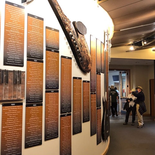 A museum exhibit features informational plaques on a curved wall and two visitors reading and taking photos.