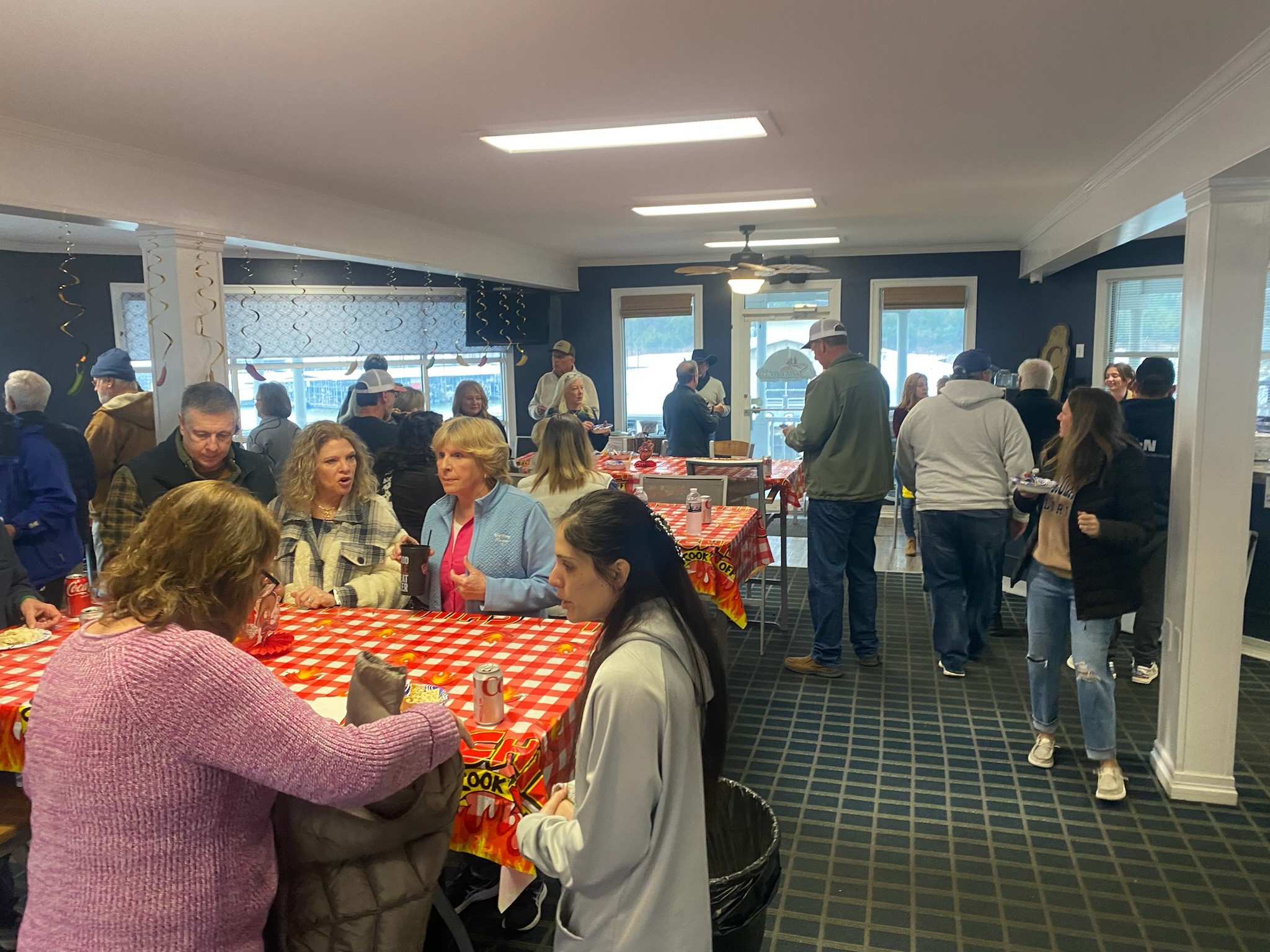A lively indoor gathering with people socializing around tables covered with red-checkered tablecloths, set in a warmly lit room with large windows and decorative streamers.