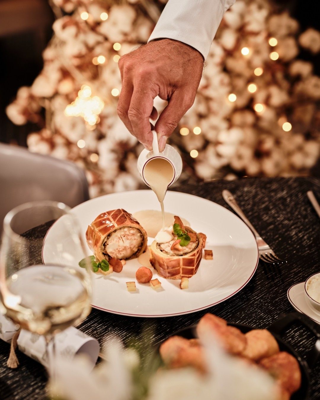 Chef pouring sauce over a gourmet Christmas entrée at Dhoom, showcasing fine dining holiday craftsmanship with warm, glowing festive lights in the background.