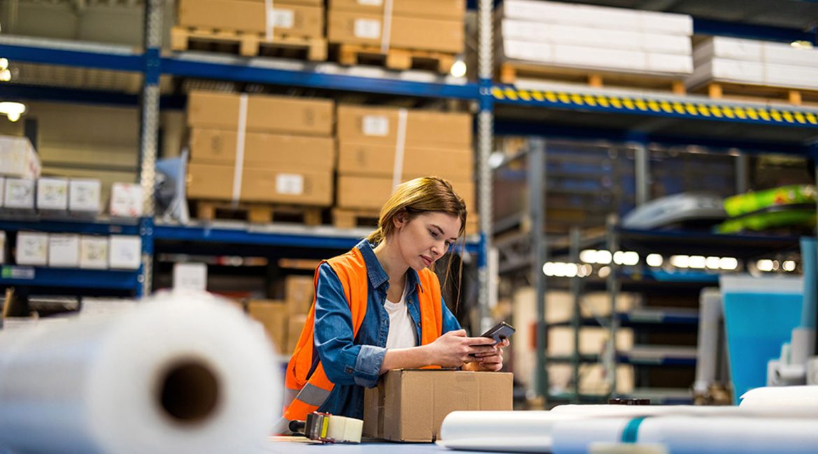 Woman in safety vest checking phone, surrounded by cardboard boxes and warehouse shelving in a distribution center.