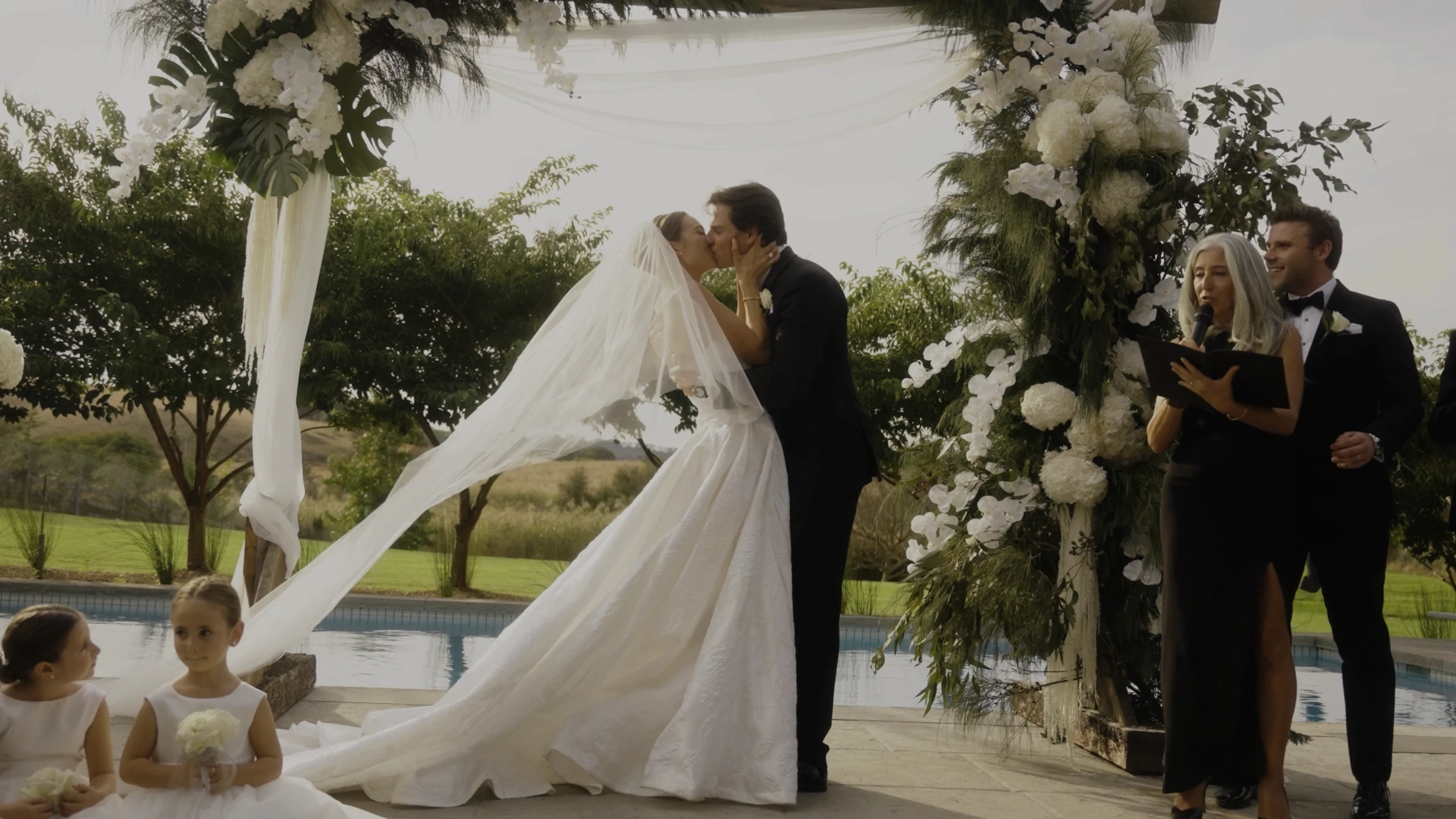 A bride and groom share a kiss under a floral arch with white roses and greenery, as bridesmaids and an officiant in elegant attire stand nearby in a picturesque outdoor setting by a pool.