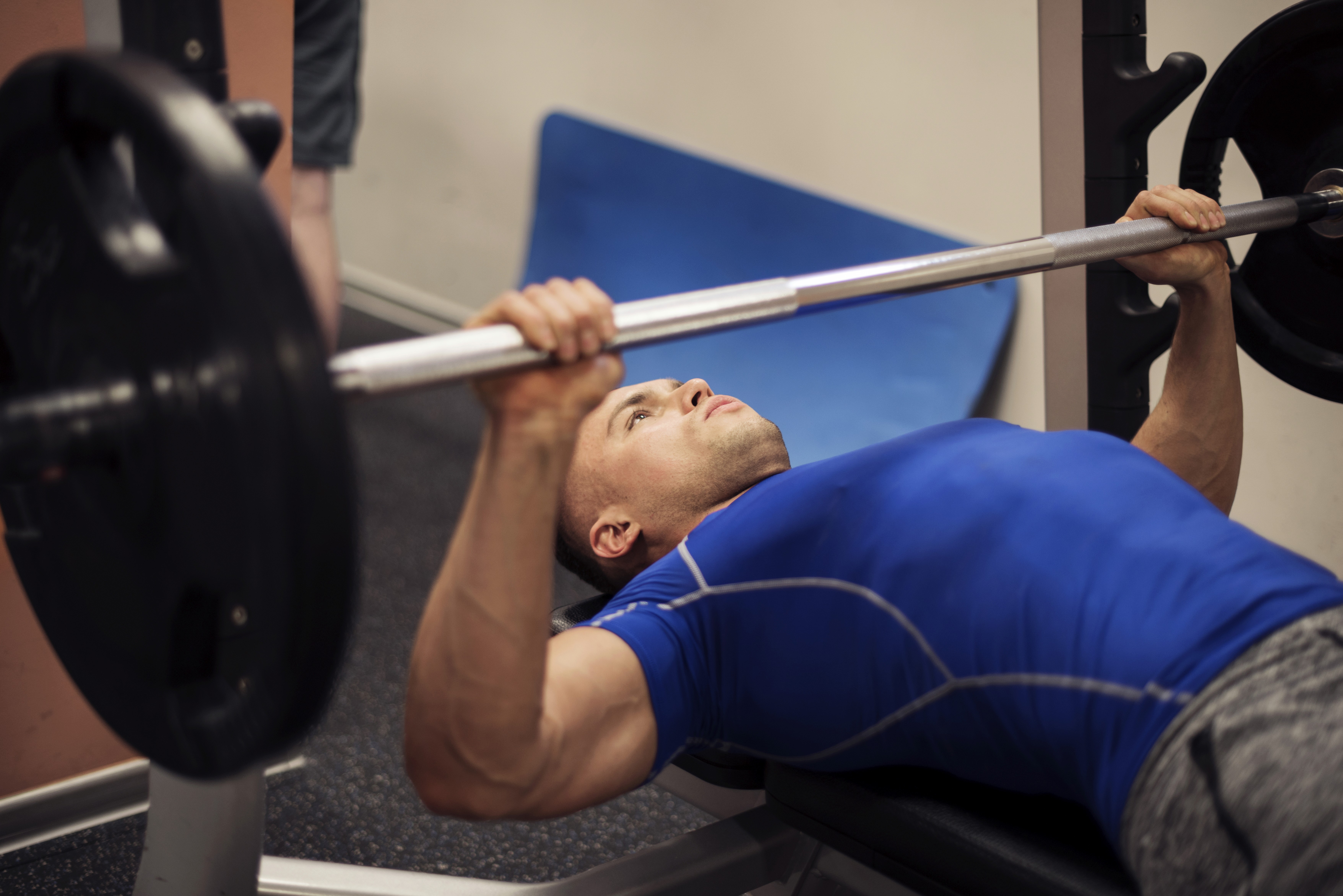 Man in a blue shirt performing a bench press with a barbell, focusing on lifting weights while lying on a bench in a gym.