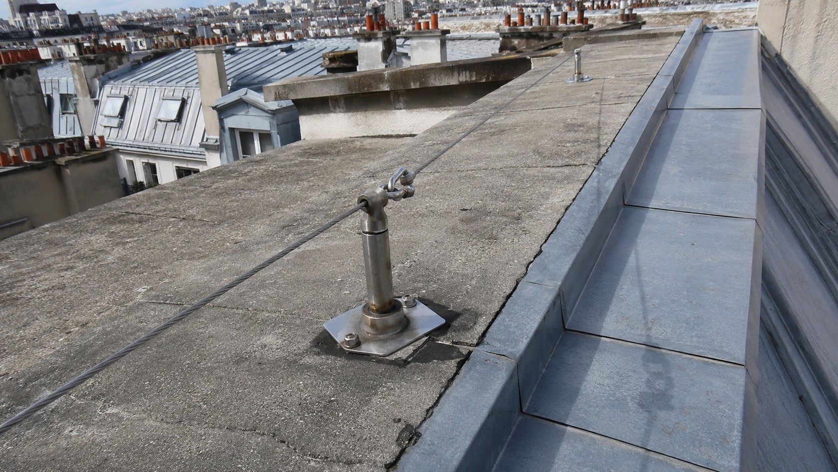 Ligne de vie en toiture d’immeuble à Paris avec câble de sécurité inox, point d’ancrage métallique et toiture zinc sous soleil.
