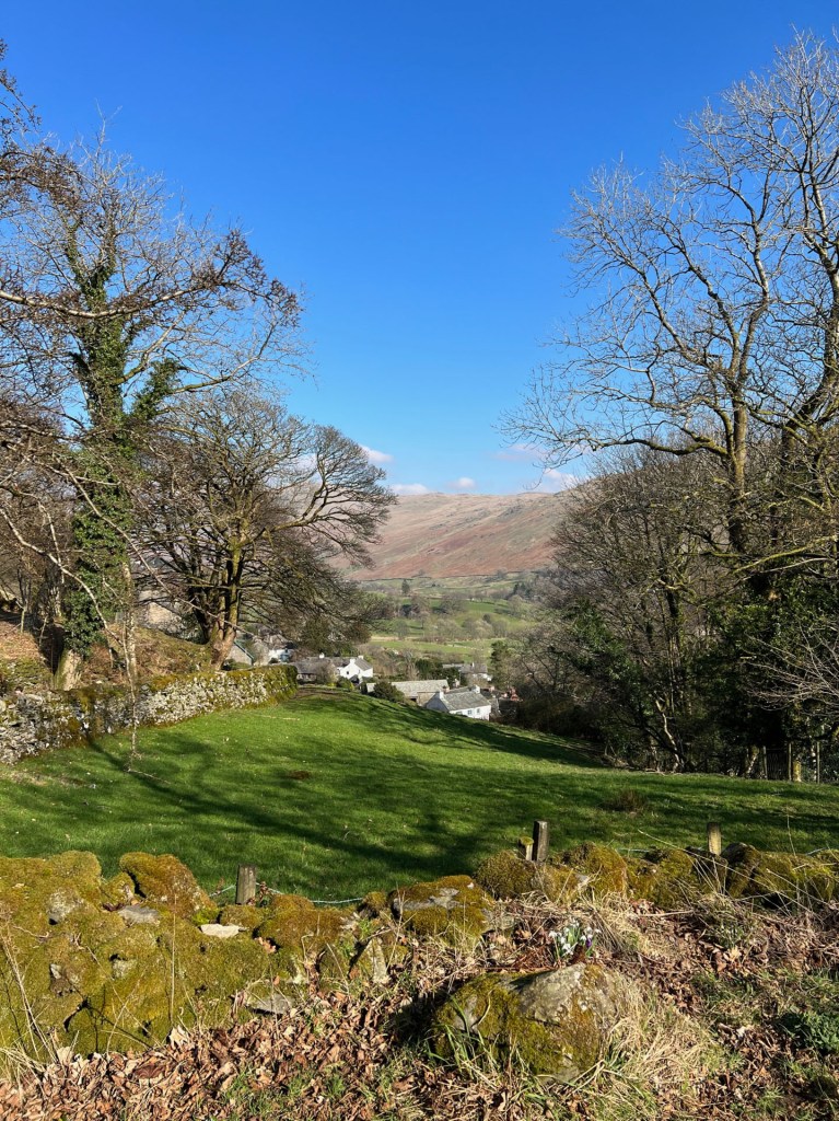 A view over a stone wall, looking down a green field that leads to the village of Troutbeck at the bottom. A few small white houses in the middle, with feels in the background and a clear blue sky.