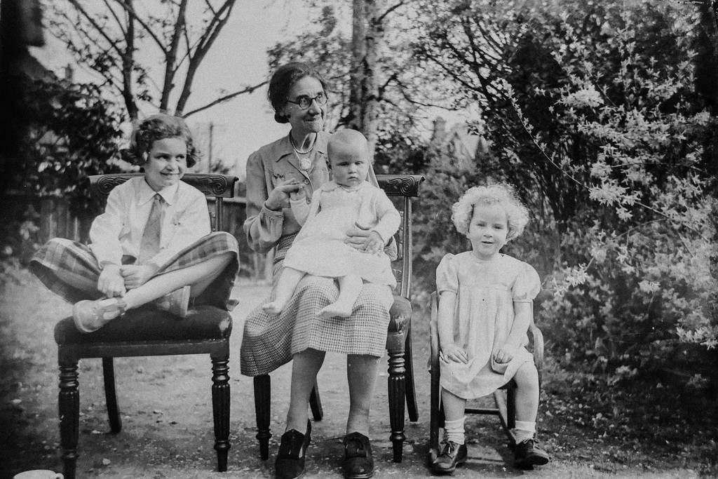 A vintage black-and-white photo features a grandmother sitting outdoors with her three young grandchildren. The grandmother holds the baby on her lap, while the other two children sit on chairs beside her. The background showcases a garden with trees and bushes, evoking a sense of nostalgia and family heritage.