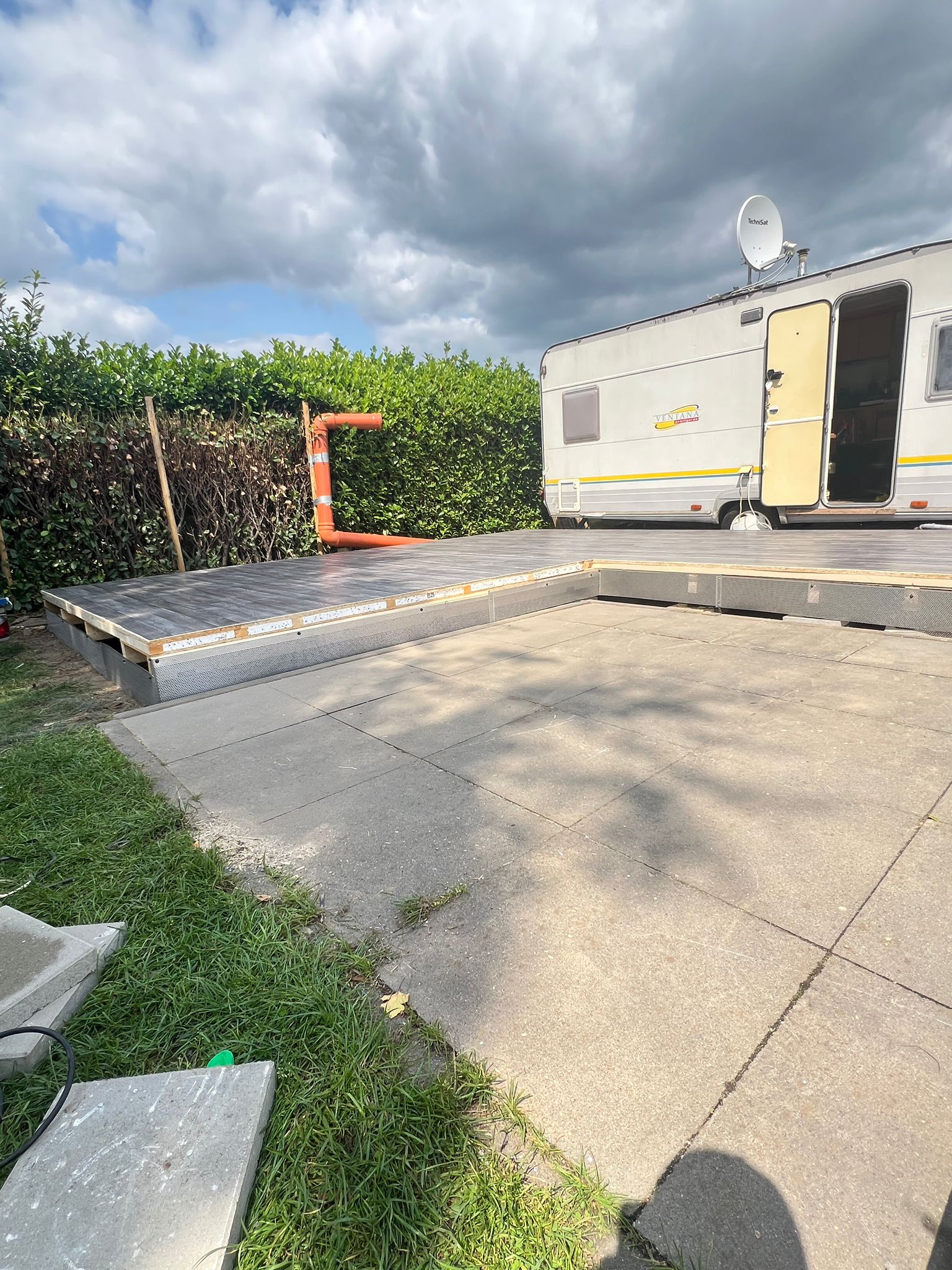 Ein Wohnmobil auf einer Terrasse mit Grün, teilweise gebautem Deck und bewölktem Himmel, mit einem Wohnmobilschutzdach und einer Terrassenverkleidung, während ein Innenausbau im Gange ist.
