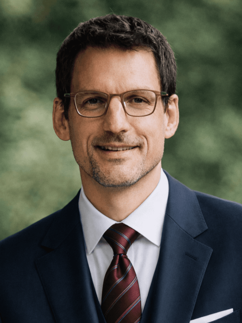 A young man with dark hair smiles at the camera, set against a modern indoor background.