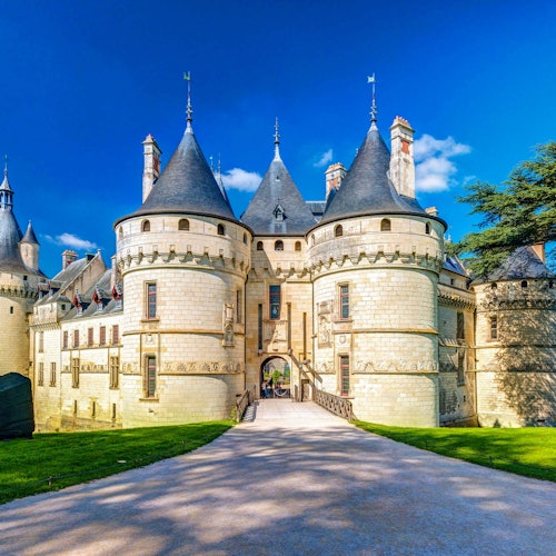 Château de pierre avec des tours coniques, une arche centrale, et un chemin pavé menant à l'entrée, sous un ciel bleu clair.
