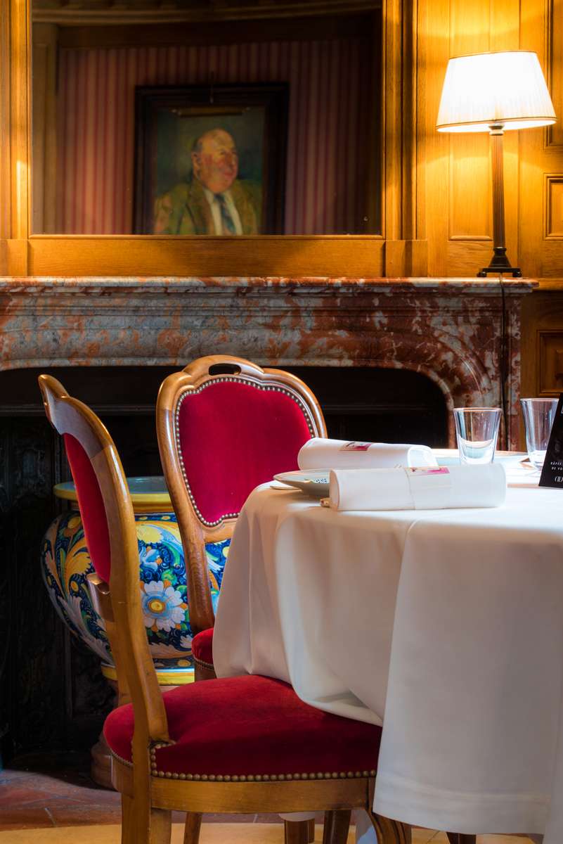 A red velvet chair and a table with a white cloth and menu sit before a fireplace with a portrait above it.