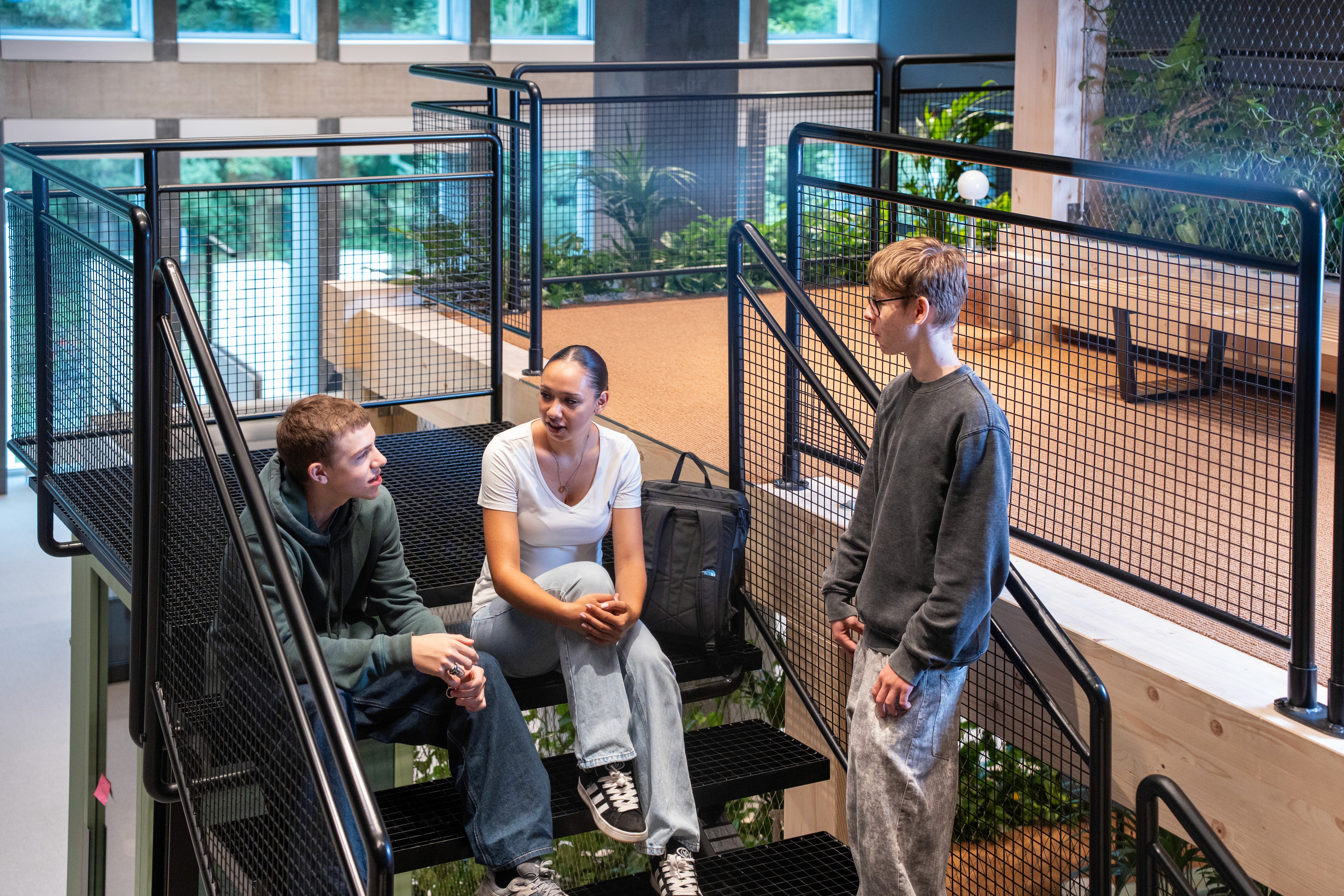 A young person with curly hair smiles at the camera while sitting in front of a laptop against a dark background.