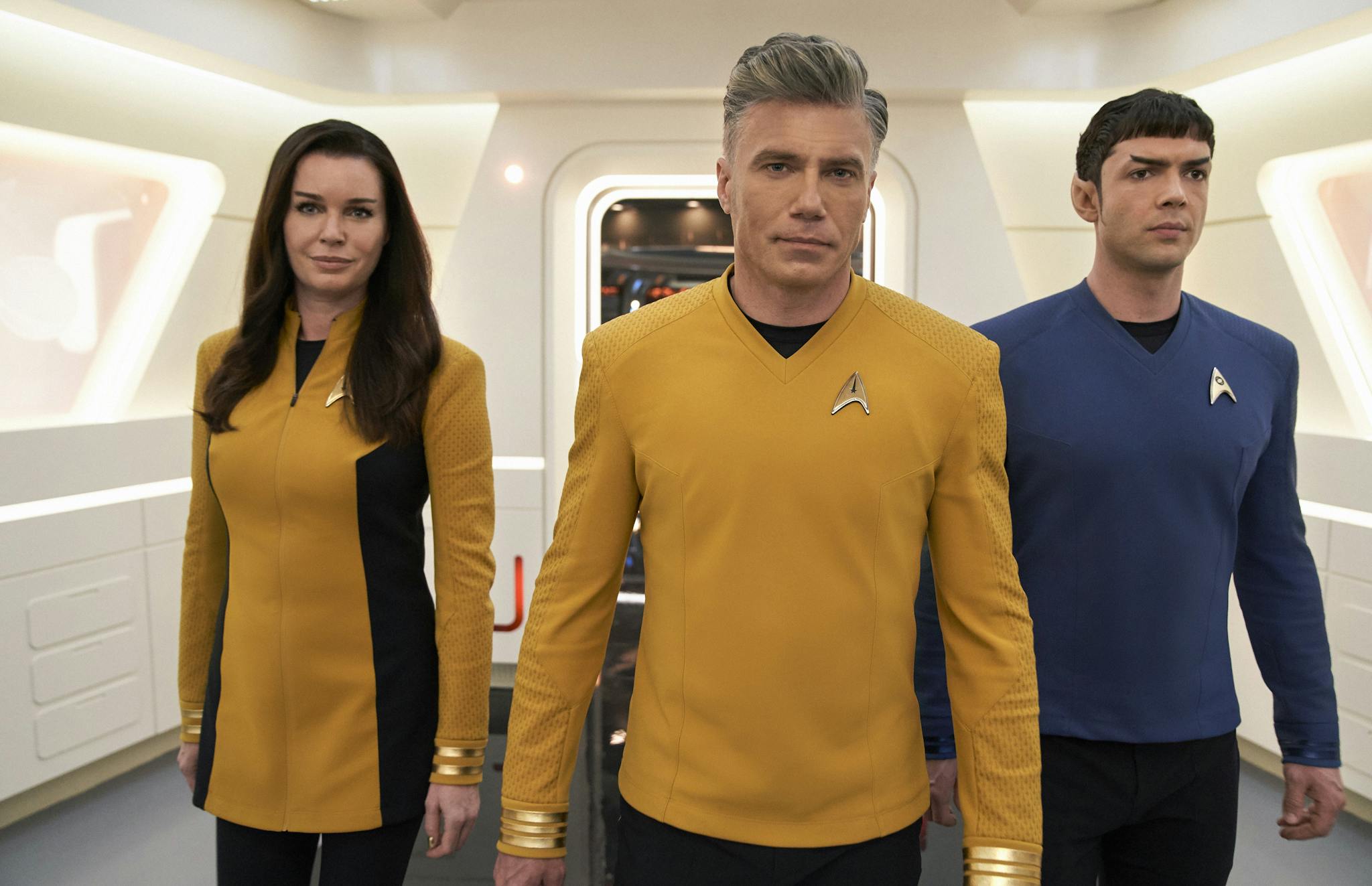 Three people in futuristic uniforms walk confidently inside a bright sci-fi spaceship interior. The two on the left wear yellow uniforms, while the person on the right wears a blue one. The setting features sleek white walls and modern lighting.