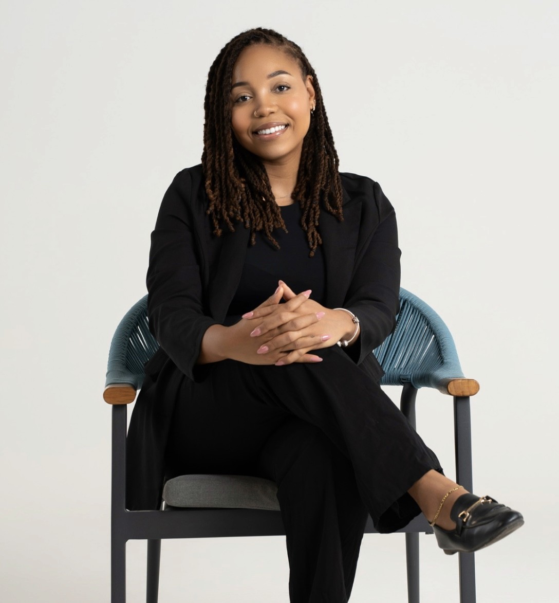 Woman in a stylish orange visor, yellow earrings, and a navy lace top against a blue backdrop. Her expression is poised, with bold red lipstick.