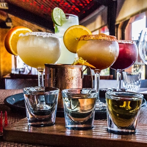 A variety of colorful cocktails and shots on a wooden board inside a dimly lit bar or restaurant.