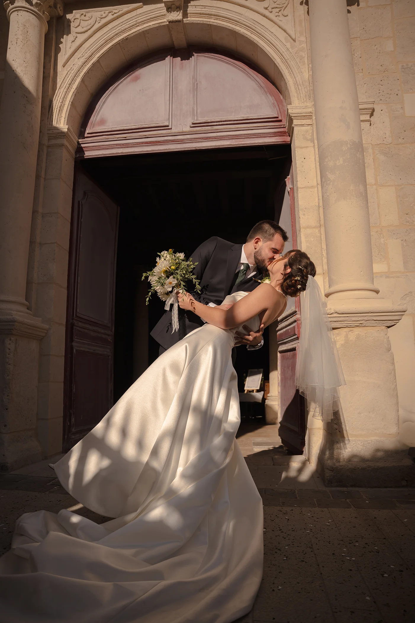 Groom dipping the bride for a kiss in front of the arched wooden doors of a stone church, her satin train spread on the ground