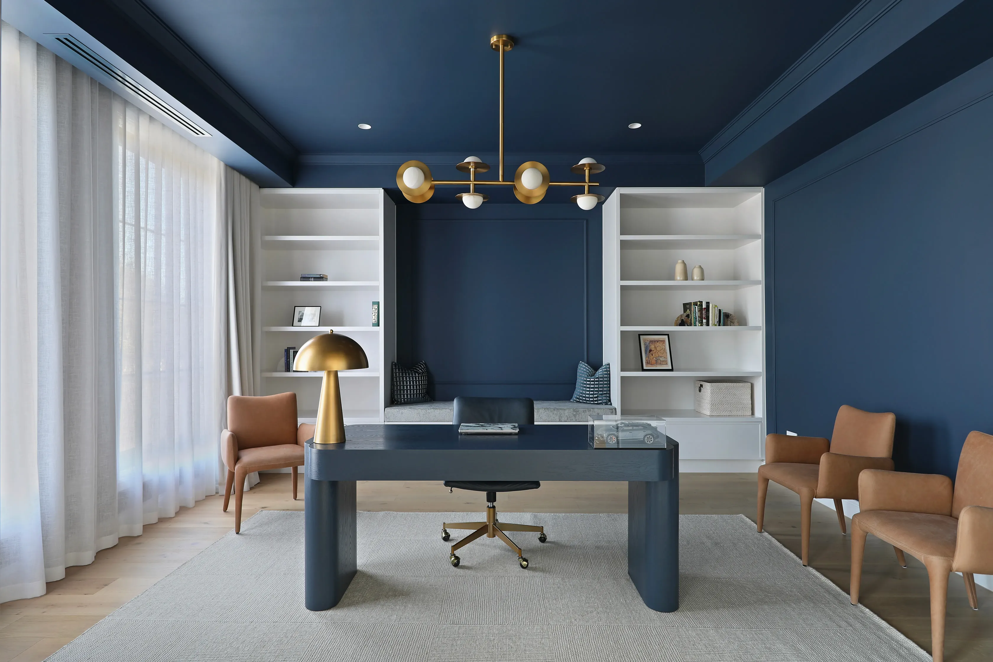 Navy-blue study with panelled walls and ceiling, white built-in shelving flanking a window seat, a brass orb chandelier, a dark timber desk with brass lamp, leather office chair and tan armchairs on a grey rug