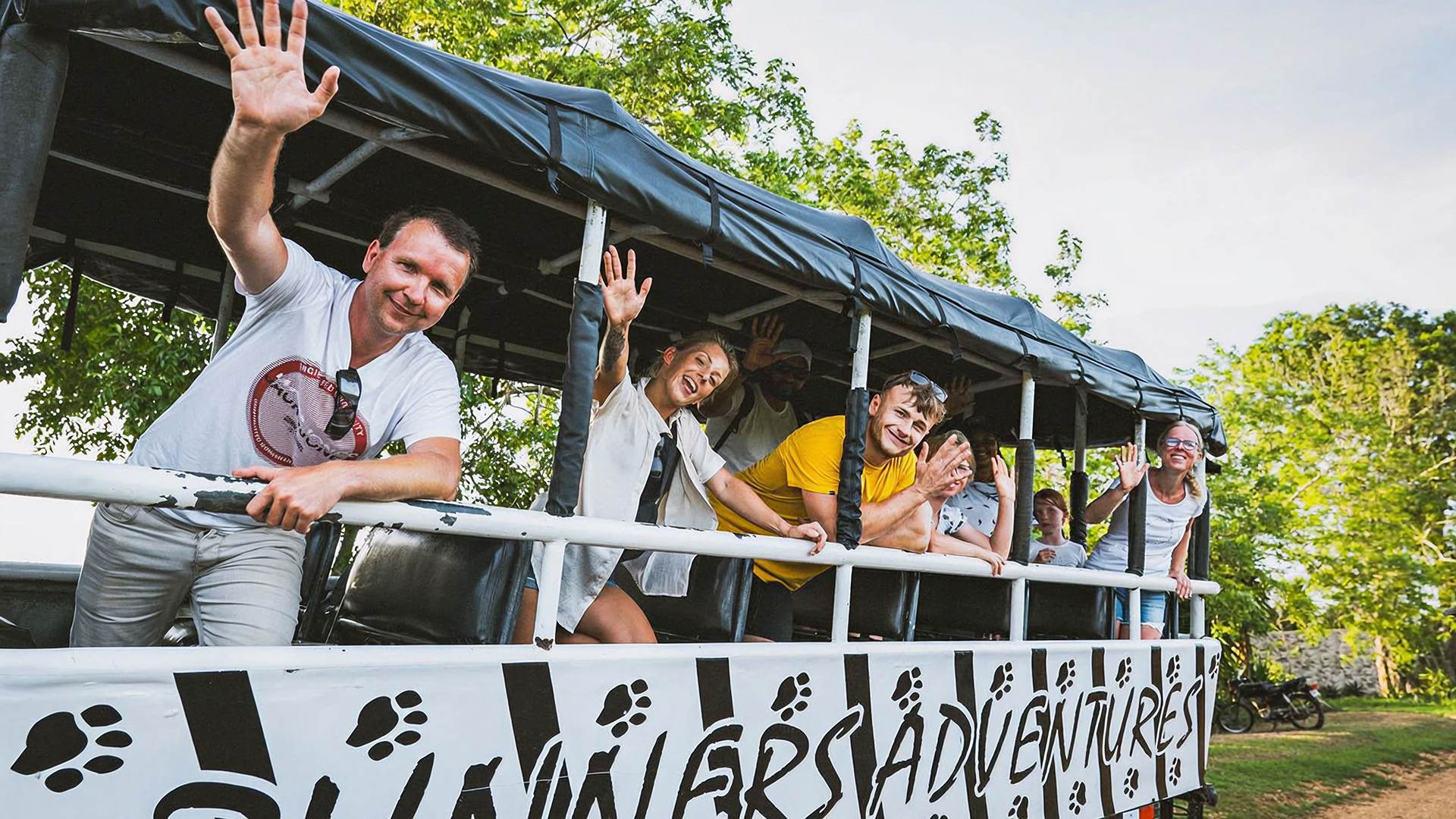 Group of smiling tourists waving from an open-air safari truck during an adventure tour.
