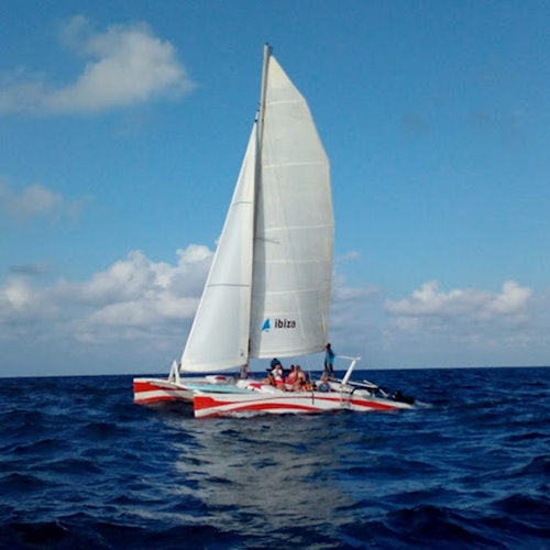 一艘紅白色船身的雙體船在藍天之下的海洋上航行，甲板上坐著一些人。