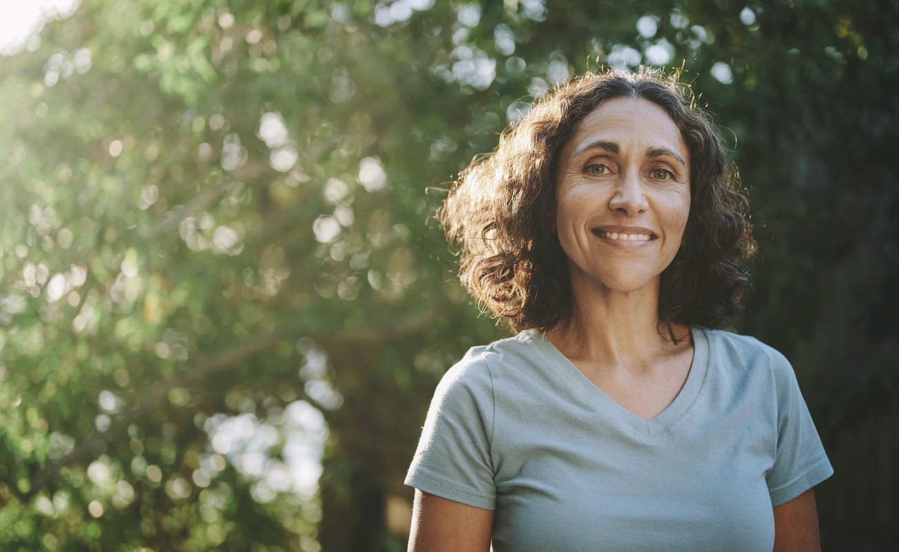 A middle-aged woman in a t-shirt smiling outside near trees